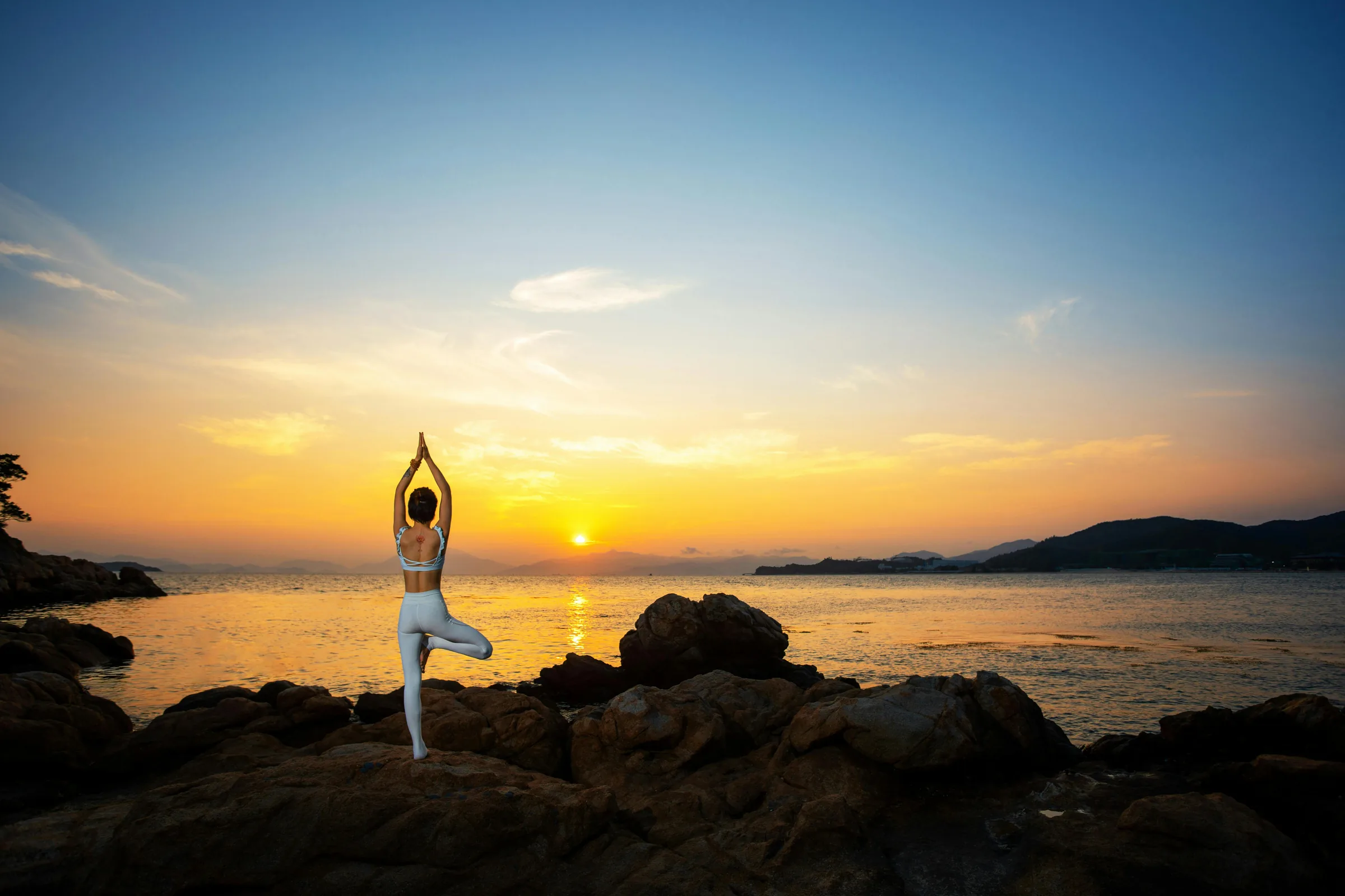 Woman in tree pose on rocks overlooking the Aegean at golden sunset.